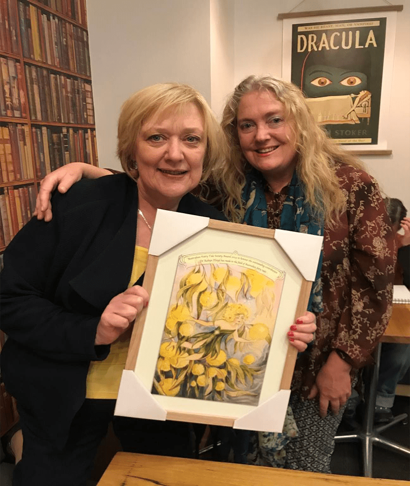 Dr Robyn Floyd shows a framed picture while Leila Honari has her arm over her shoulders, in a room with a bookcase and a comical Dracula poster.