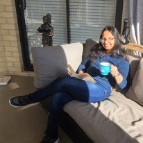 Devahuti Rai Chaliha sits on a sofa smiling and holding a mug. Behind her is an Indian statue.