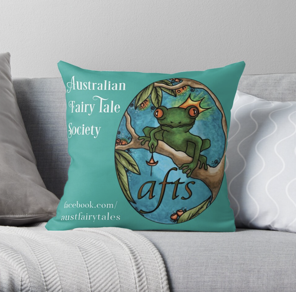 Bright green cushion sits on grey couch. The cushion is printed with the Australian Fairy Tale Society title and Facebook address, and features the AFTS logo being a crowned frog on a tree branch.