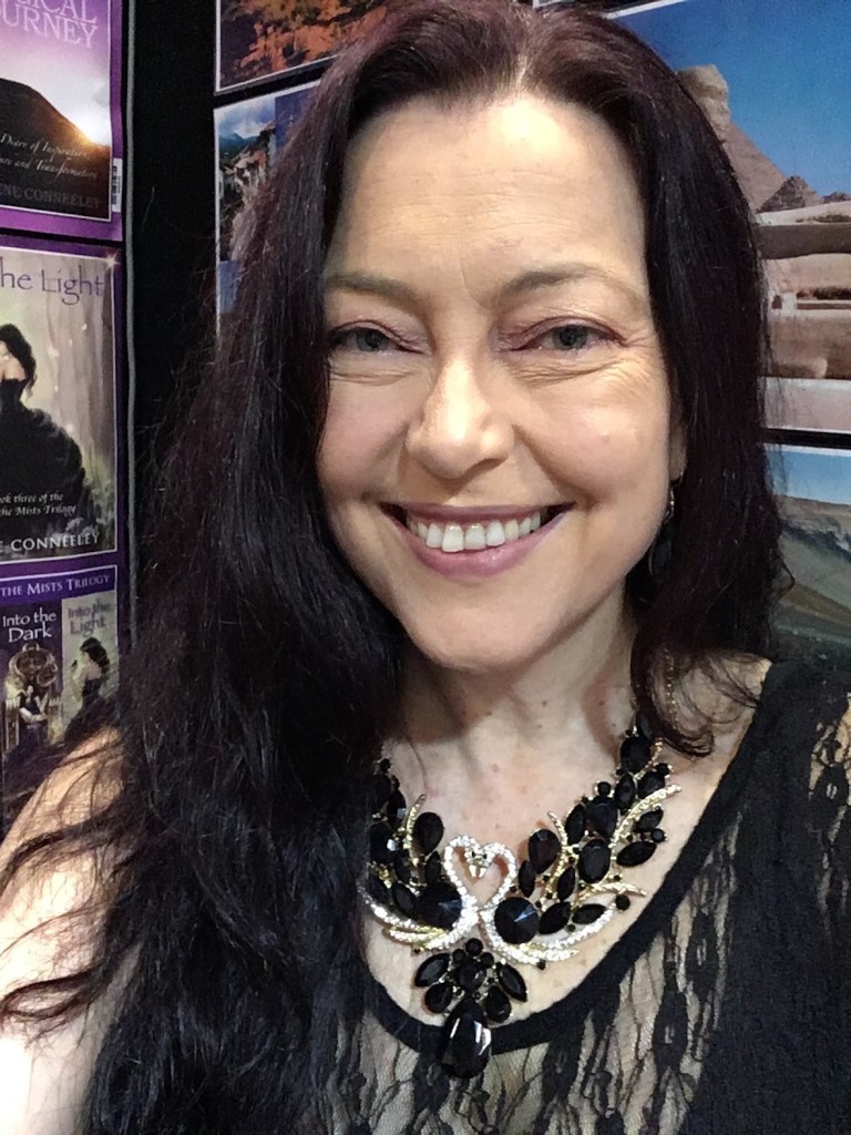 Serene Conneeley smiles in front of posters of her books and travel posters. She wears a dark crystal necklace with light crystals at the centre forming the shape of two swans making a heart shape as they touch heads.