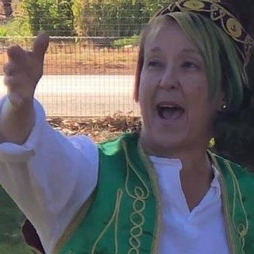 Jo Henwood raises her hand as she sings or shouts, wearing an embroidered bolero and cap in front of a tree and a fence which could be school grounds.
