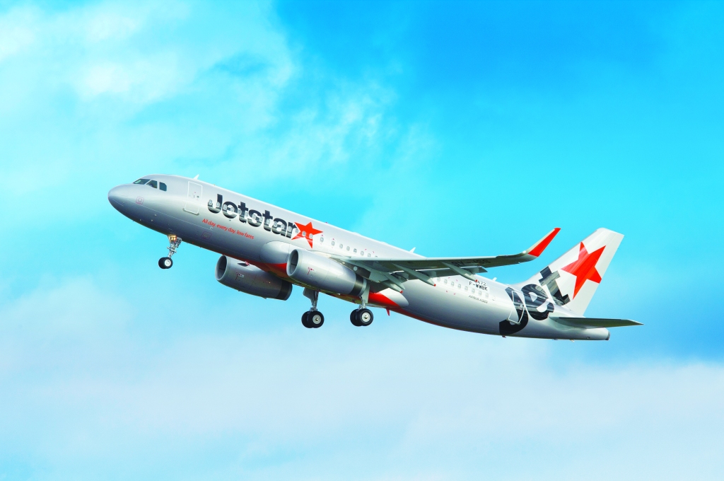A 2-engine passenger jet with Jetstar logos flies up into a bright blue sky.