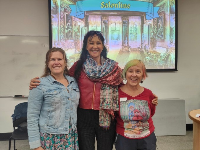 Three smiling women stand in front of a screen showing a salon with ornate furnishings with a banner saying Salonline above it. The women are in colourful casual clothes, with Alyssa Curtayne on the left, Eliane Morel in the middle, and Jo Henwood on the right.